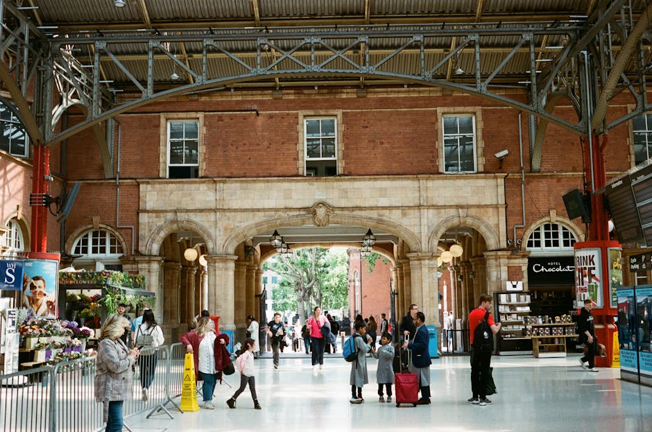 Inside a bustling train station with a high, arched metal ceiling supported by large steel beams and red structural columns, a variety of people are gathered. Some individuals are waiting, chatting, or walking, with a few carrying luggage or backpacks. At the edge of the station, there is a flower stall with fresh bouquets and potted plants, protected by metal barriers, and a small shop selling chocolates and snacks, with shelves and signage visible. Several people are holding or examining documents, possibly tickets or travel papers, while others are engaged in conversations or looking at their phones. Natural light filters through large windows on the brick walls above, illuminating the scene. On the platform, there are loading and unloading areas with some individuals lifting or moving boxes, wrapped in cardboard or plastic, towards a waiting vehicle or storage space. This scene reflects a busy environment typical of a UK railway station involved in home relocation or moving logistics, with clear indications of packing and furniture transport activities, consistent with professional removals services occasionally seen in case of house moves.